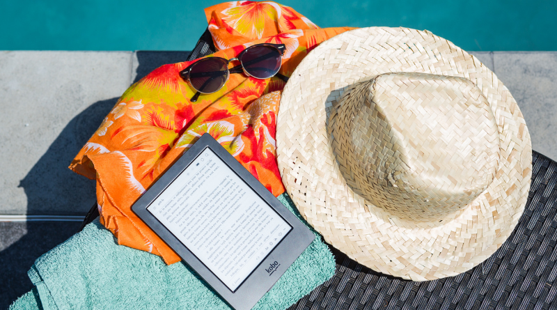 eBook reader on top of a pile of beach clothes, hat, towel and sunglasses